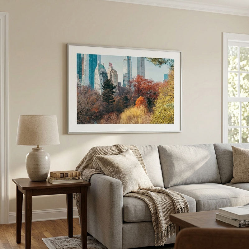 A framed art print of a city skyline with autumn trees is displayed on a beige wall above a gray sofa in a styled living room setting.