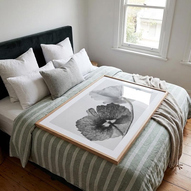 A large framed black and white botanical art print with a poppy design, resting on a styled bed in a bright, modern bedroom.