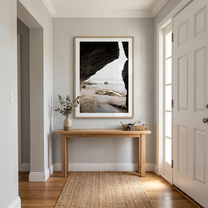 A framed black and white coastal rock print hangs on a light gray wall above a wooden console table in a bright entryway with hardwood floors and a woven rug.