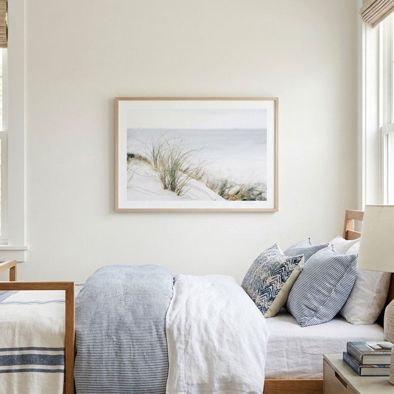 A framed art print depicting a serene beach dune landscape with sand grasses, hung on a light cream wall above a neatly made bed with blue and white striped pillows and bedding in a bright, modern bedroom.