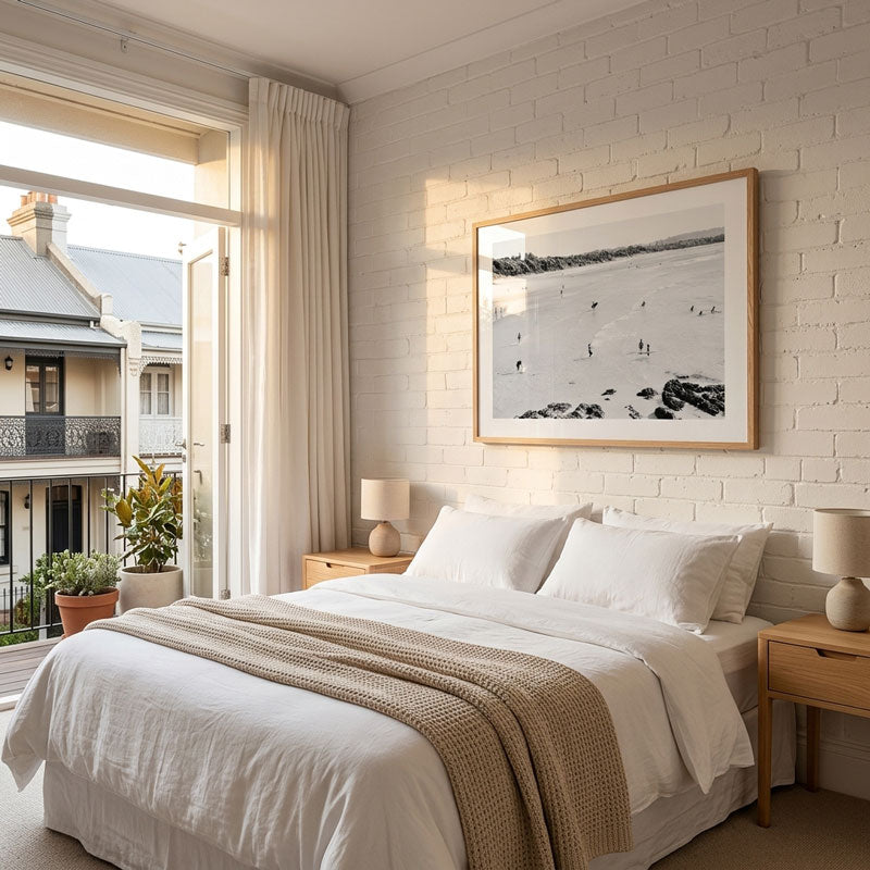 A styled bedroom interior with a neatly made bed, white bedding, and a beige throw blanket, set against a white brick wall with a large framed black-and-white landscape photograph. An open window and balcony view are visible on the left.
