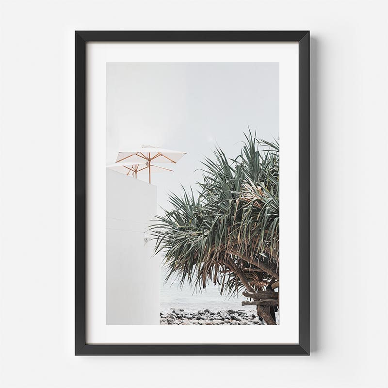 Framed artwork of a beach scene with palm trees and an umbrella on a white wall.