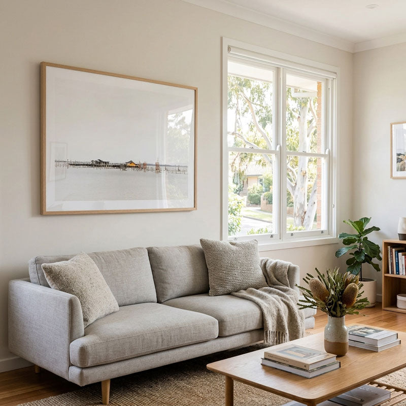A large framed minimalist art print with a light wood frame hangs on a beige wall above a light gray fabric sofa in a bright, modern living room.
