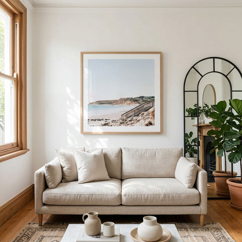 A framed coastal landscape print hangs on a white wall above a light beige sofa in a bright, modern living room with wood floors, a window, and potted plants.