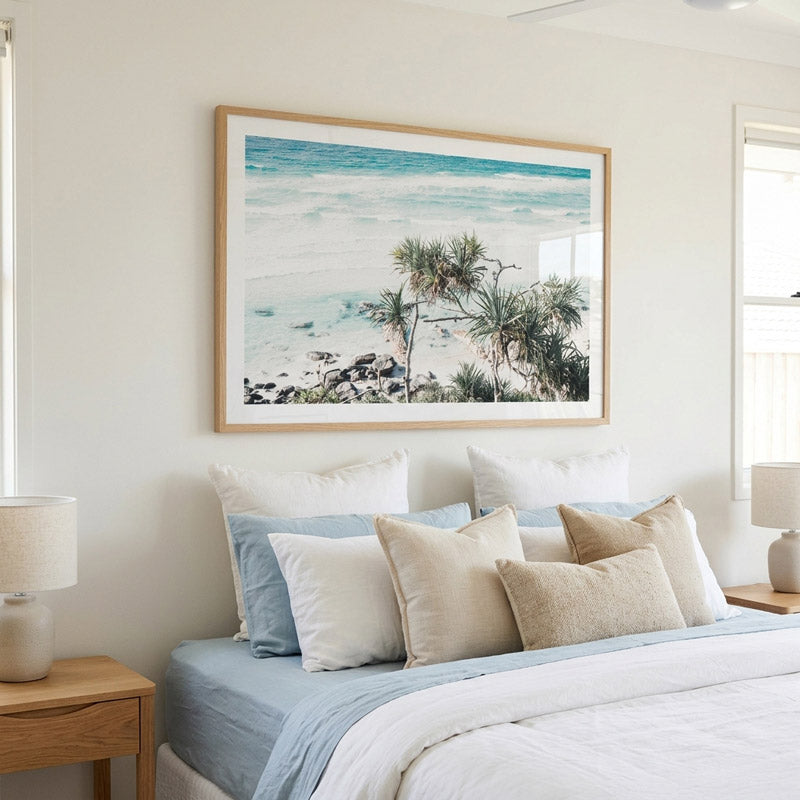 A framed beach print with turquoise ocean water, white sand, and palm trees, displayed on a light beige wall above a neatly made bed with white and light blue bedding.