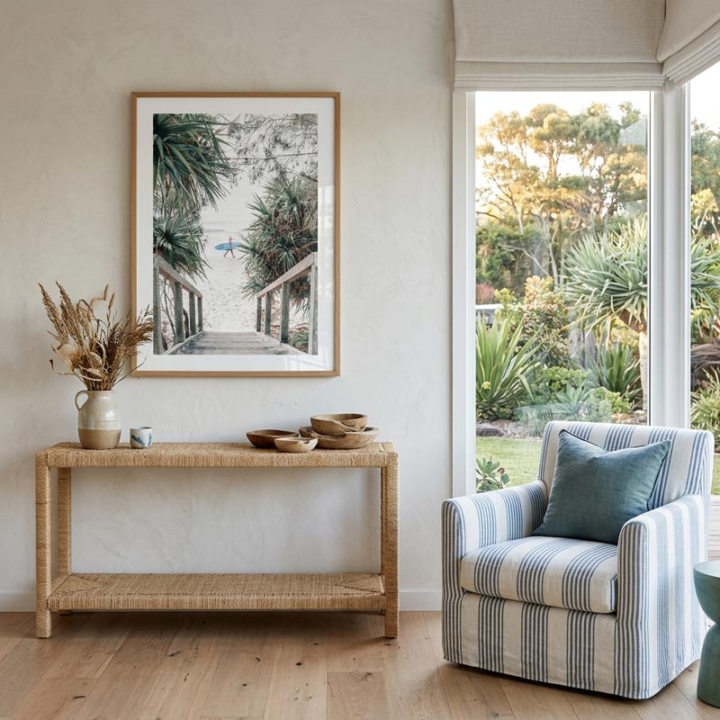 A framed art print depicting a tropical beach scene with palm trees and a wooden boardwalk, styled in a bright, modern room above a woven console table next to a blue-striped armchair.