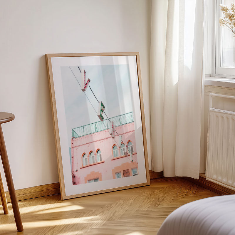 Framed artwork of a pink building on a wooden floor with a window and radiator in the background.