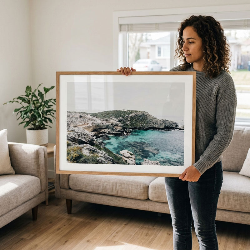 A woman stands in a bright living room holding a large framed landscape photograph of a rocky coastline and turquoise bay. The print has a white mat and a light wood frame, and the room has a beige sofa, wood floor, and a potted plant in the background.