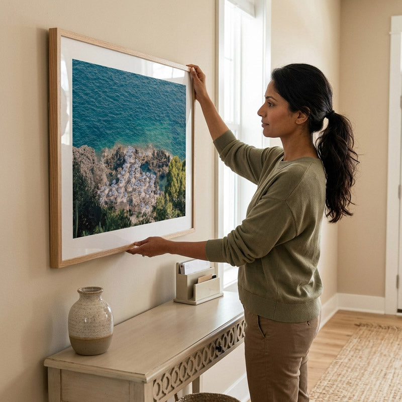 A woman in an olive green sweater and khaki pants hangs a large framed landscape photograph of a rocky coastline with blue water on a beige wall in a bright room.