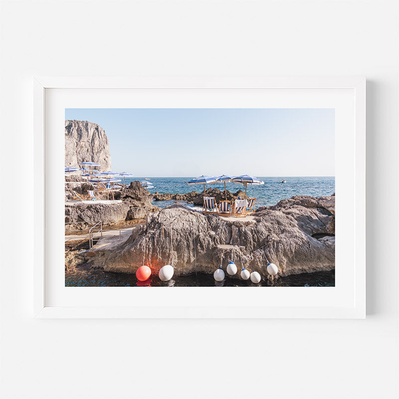 Framed photograph of a rocky coastal scene with chairs and umbrellas.