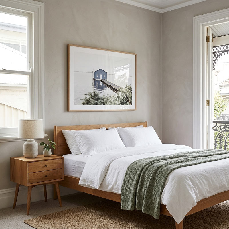 A styled bedroom scene featuring a wooden bed frame with white linens and a green throw blanket, next to a matching wooden nightstand with a lamp. A framed black and white architectural print hangs on the textured beige wall above the bed.