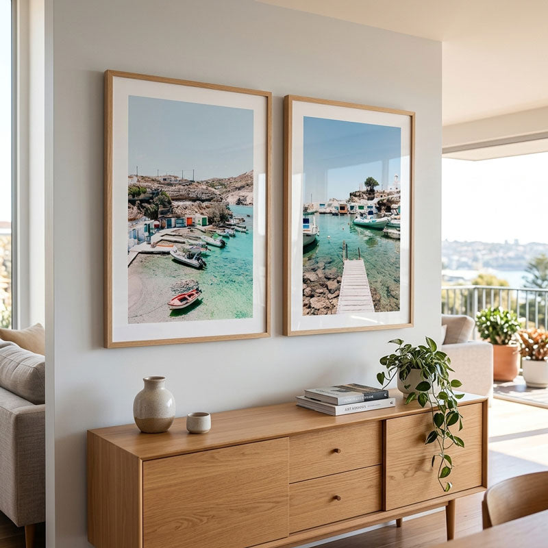 Two framed coastal harbor prints with light wood frames hang on a light gray wall above a wooden console table in a bright modern living space.