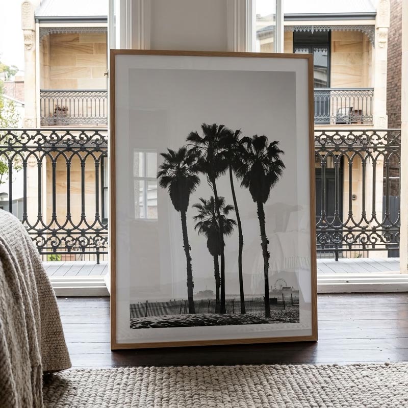 A framed black and white art print of palm trees, leaning against a window in a modern room with a wooden floor and a woven rug.