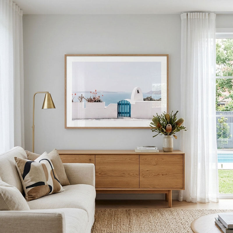 A framed art print depicting a Mediterranean coastal scene with white buildings and a blue bench, displayed on a light gray wall above a mid-century modern wooden sideboard in a bright, contemporary living room.