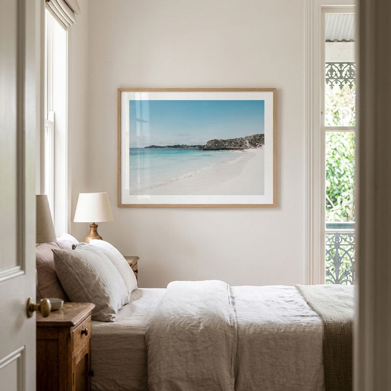 A framed beach landscape print hangs on a light beige wall above a neatly made bed in a softly lit bedroom. The artwork shows a calm turquoise ocean, white sand, and rocky shoreline, with a light wood frame and white mat.