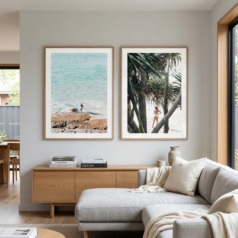 Two framed beach-themed art prints hanging side-by-side on a light gray wall above a wooden console and a modern light gray sectional sofa.