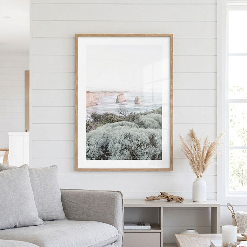 A framed coastal landscape art print hangs on a white shiplap wall above a light gray sofa in a bright, modern living room. The print depicts sea stacks and coastal vegetation, framed in light wood with a white mat.