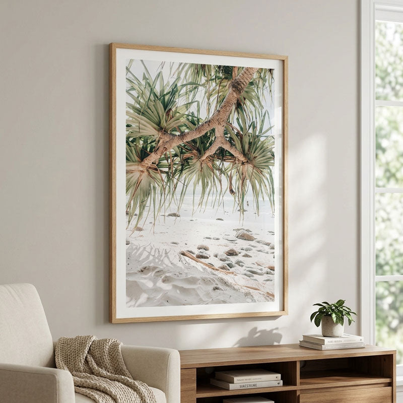 A framed vertical art print depicting green palm-like foliage over a sandy beach, hanging on a light gray wall above a wooden console table in a styled living room.
