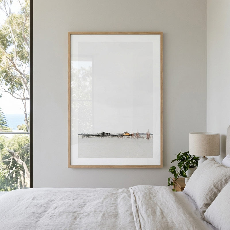 A minimalist framed art print featuring a pier over calm water, styled on a light gray wall above a bed with white linens in a bright, modern bedroom.