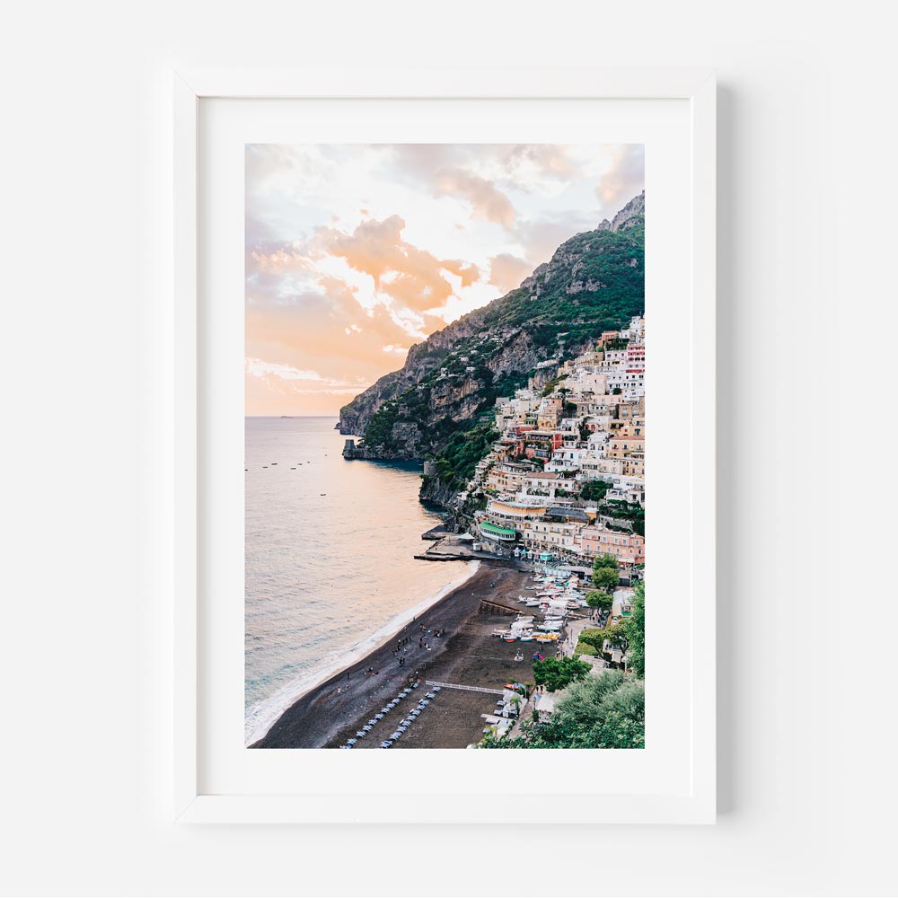 A framed wall art featuring a coastal town perched on a cliffside by the ocean, with a colorful sky above.