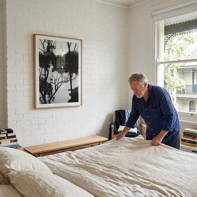 A man in a navy blue shirt is adjusting a textured cream-colored duvet on a bed in a bright, modern bedroom with white brick walls and a framed black-and-white art print.