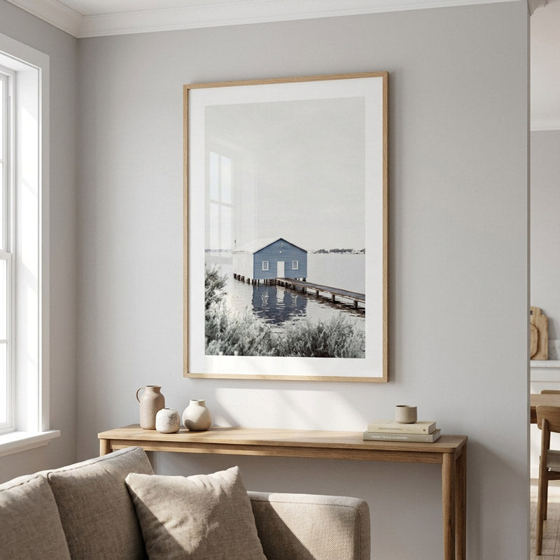 A framed black and white coastal art print featuring a small blue house reflected in water, displayed on a light gray wall above a wooden console table in a modern living room setting.