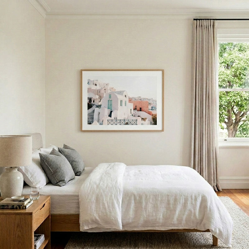 A framed art print depicting a Mediterranean-style coastal town with white and terracotta buildings, hung on a beige wall above a neatly made bed in a modern, softly lit bedroom.