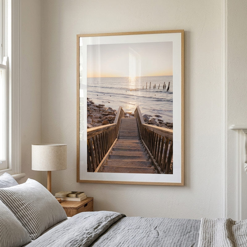 A framed vertical art print of a wooden boardwalk leading to a rocky seaside at sunset, displayed on a bedroom wall above a bed with gray bedding and a bedside lamp.