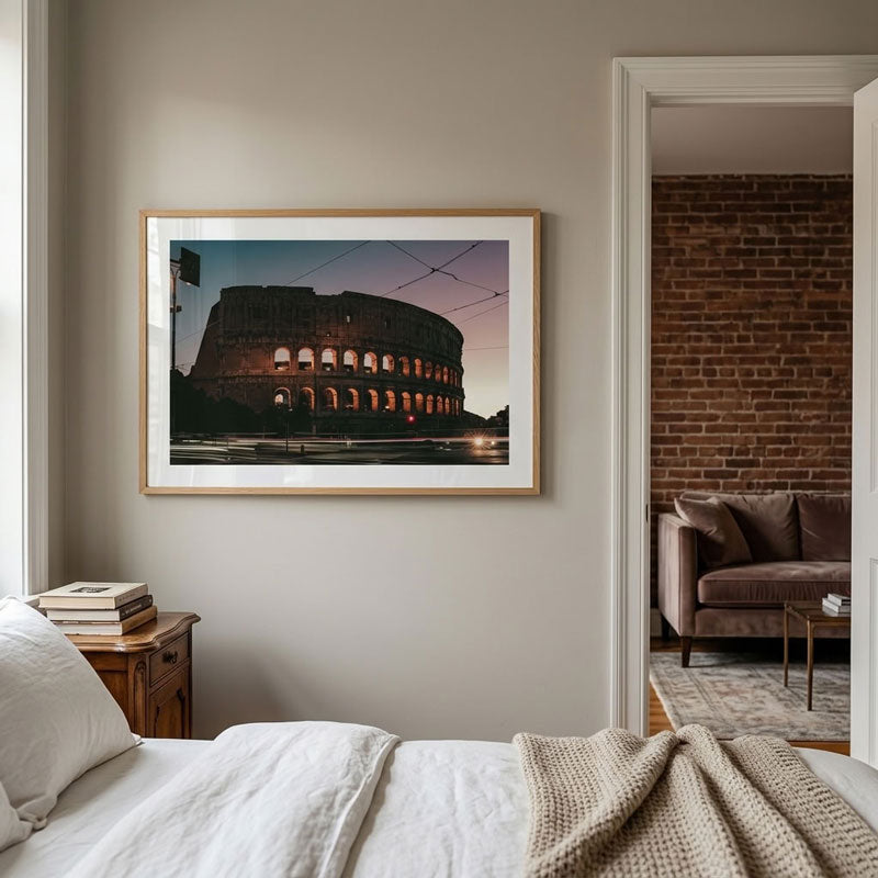 A framed art print of the Colosseum in Rome at dusk, displayed on a neutral beige wall in a modern bedroom setting.