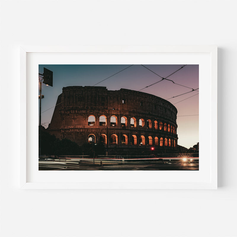A framed photographic print of the Colosseum in Rome at dusk, shown against a clean white wall. The print features the ancient amphitheater illuminated against a twilight sky, enclosed in a simple white frame.