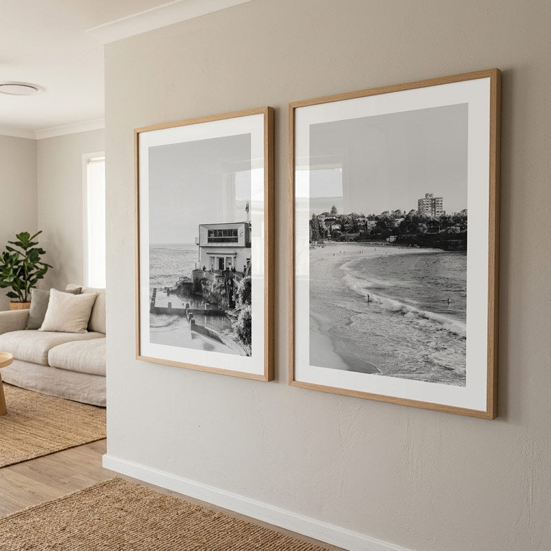 Two framed black and white beach and coastal architecture photographs with light wood frames, displayed side-by-side on a beige wall in a modern living room setting.