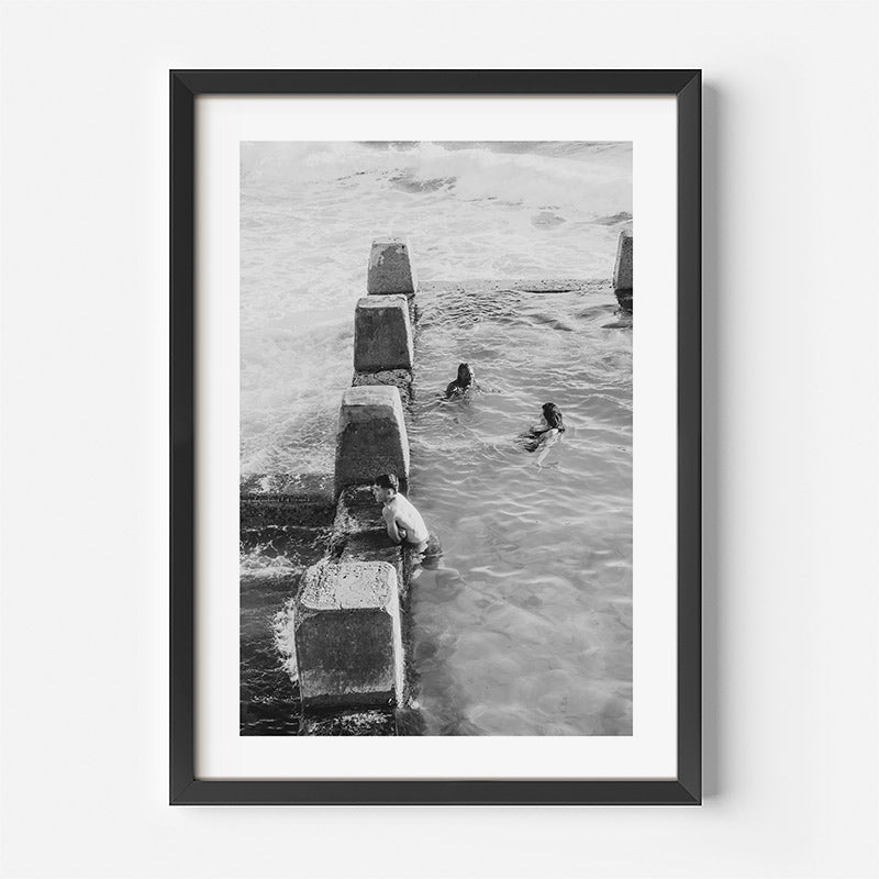 Framed black and white photograph of people swimming between concrete blocks in water.