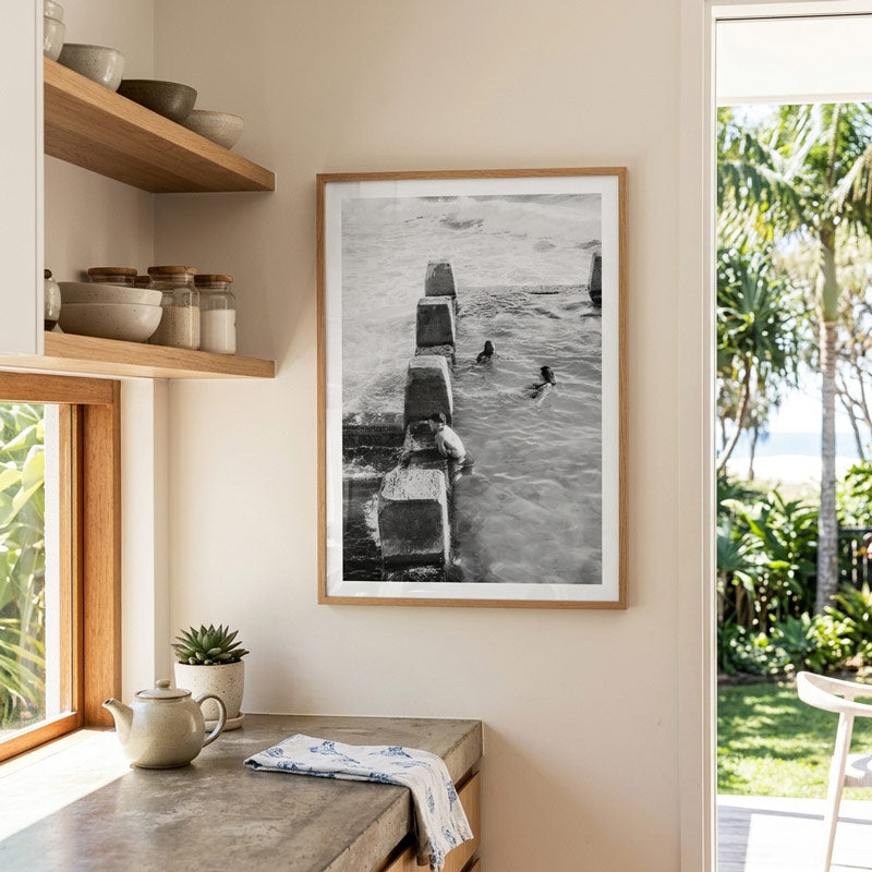 A framed black and white photograph of a pier scene with surfers hangs on a beige wall in a modern kitchen setting with wooden shelves, a countertop, and an outdoor view.