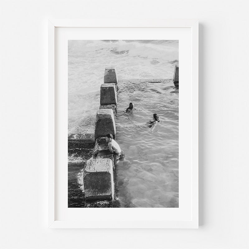 Framed black and white photograph of people swimming near concrete blocks in water.