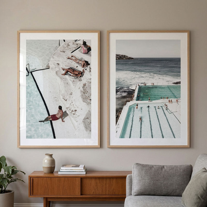 Two framed photographic prints hang side-by-side on a beige wall above a mid-century modern wooden console table. The left print shows an aerial view of a beach with people sunbathing by a pool, and the right print shows an aerial view of an outdoor swimming pool next to the ocean.