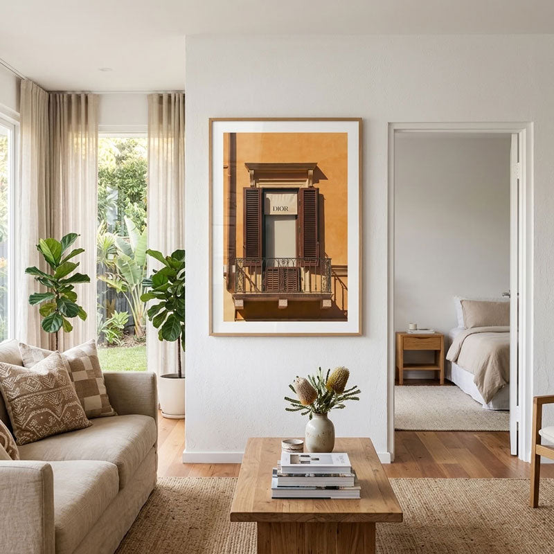 A framed art print depicting a warm-toned balcony with brown shutters, displayed on a white wall in a modern, sunlit living room with a beige sofa and wooden coffee table.