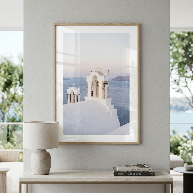 A framed art print depicting a white church on a cliff overlooking a blue sea, hung on a light gray wall above a console table with a lamp and books.