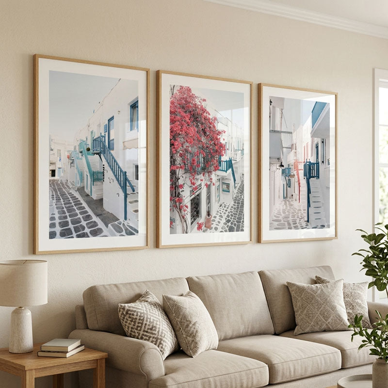 Three framed art prints depicting Mediterranean street scenes with white buildings, cobblestone paths, and blue accents, displayed on a beige wall above a light beige sofa in a living room setting.