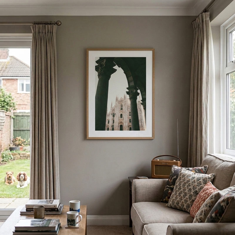 A framed art print depicting a gothic cathedral seen through dark arches, hung on a gray wall in a styled living room with a sofa, curtains, and a window showing dogs outside.