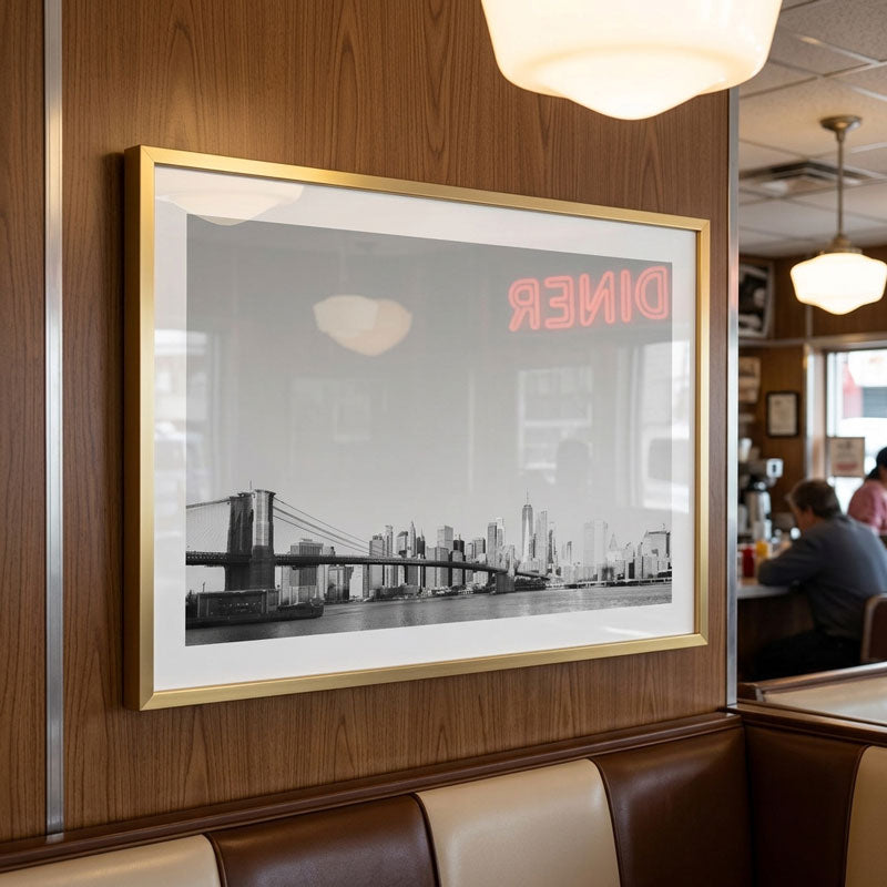 A framed black and white New York City skyline print featuring the Brooklyn Bridge and a neon-style red 'DINER' sign, displayed on a wood-paneled wall in a restaurant booth setting.