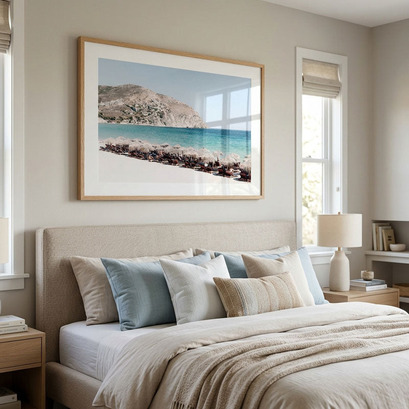 A framed beach landscape print hangs above a neatly made bed in a softly styled bedroom. The artwork shows a rocky shoreline and turquoise water, while the room features neutral beige bedding, pillows, walls, and wood furniture.