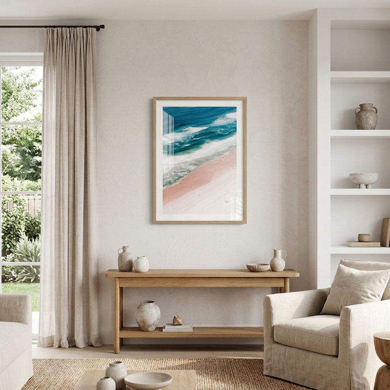 A framed vertical art print depicting a beach scene with turquoise ocean waves and a pink sandbar, displayed on a beige wall in a modern, sunlit living room above a wooden console table.