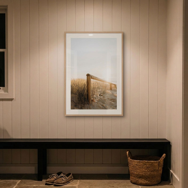 A framed vertical art print depicting a beach dune scene with a wooden fence and dry grass, displayed on a beige paneled wall above a dark bench with shoes and a woven basket below.