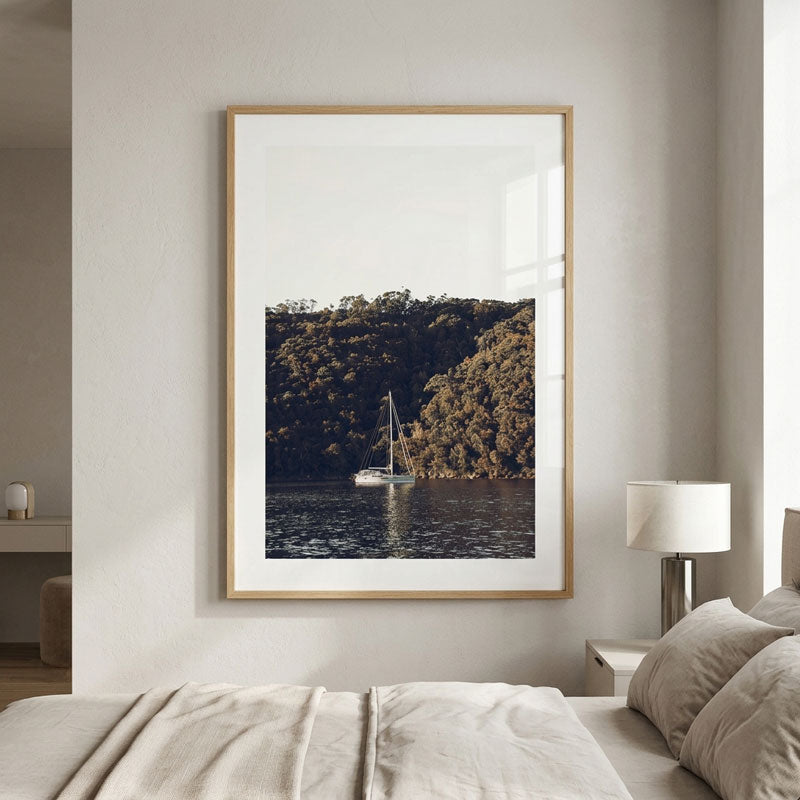 A framed vertical art print of a sailboat on a lake surrounded by a forested hillside, displayed on a light gray wall above a bed in a modern bedroom setting.