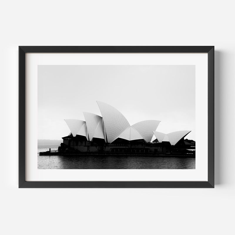 A framed black and white photographic print of the Sydney Opera House, shown hanging on a light gray wall. The print features a high-contrast monochrome image of the iconic opera house against a pale sky, mapped onto a white mat and a sleek black frame.