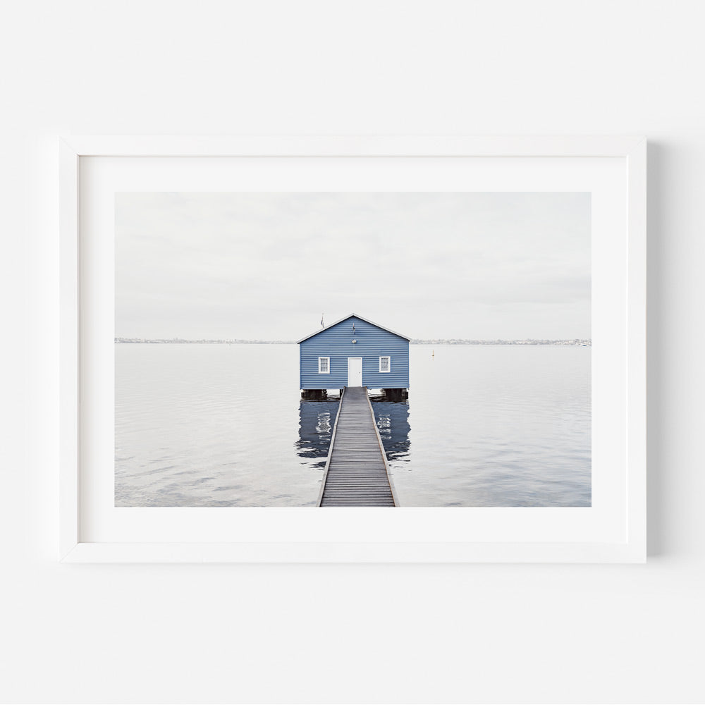 A framed photograph of a blue boat house with a jetty extending into calm water, with a predominantly white and blue color scheme.