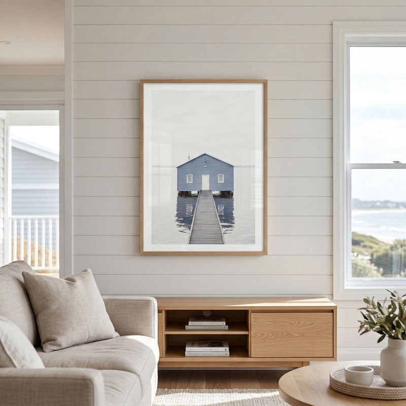 A framed art print of a blue house on a pier, displayed on a white shiplap wall above a wooden console table in a modern living room setting.