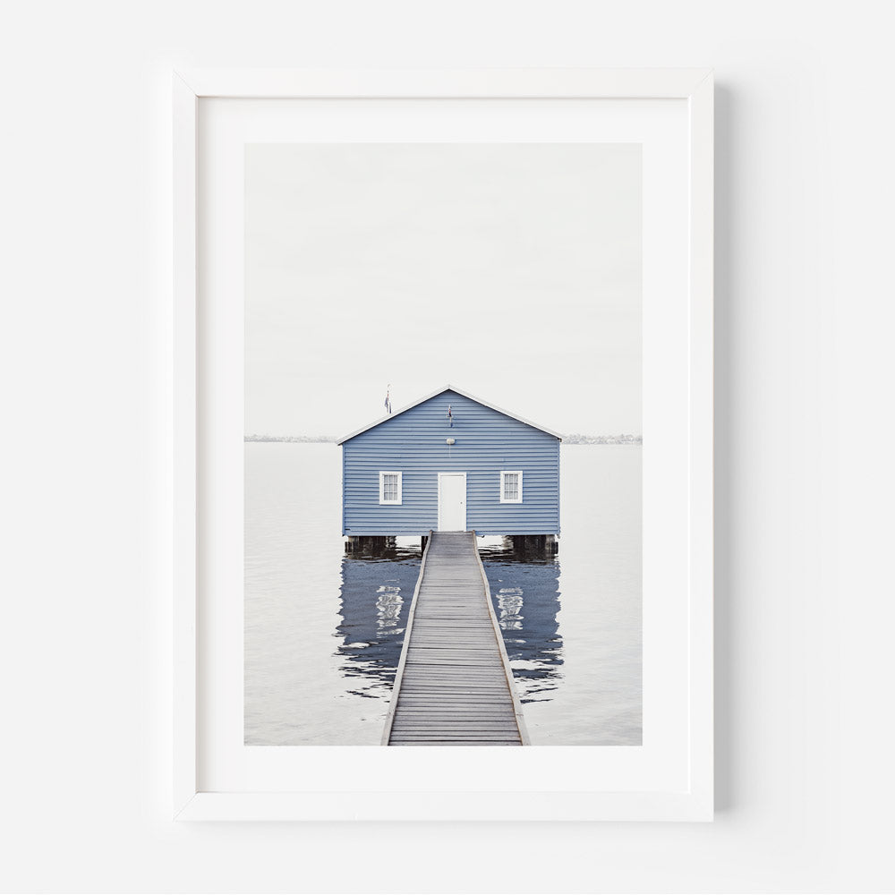 A framed photograph of a blue boat shed with a jetty leading up to it, taken from a distance, with a white border around the frame.