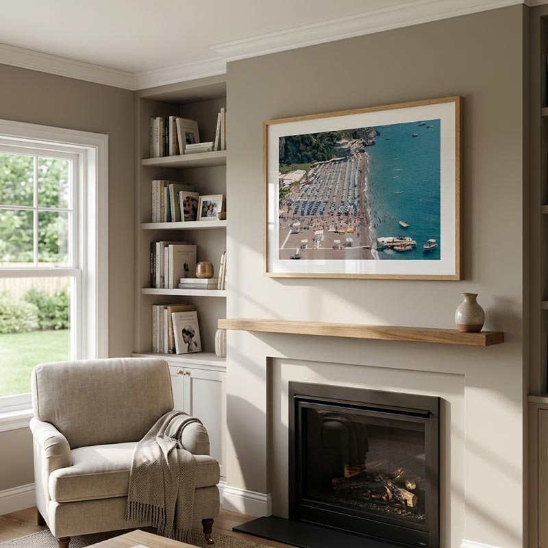 A framed art print depicting an aerial view of a coastal town and beach is displayed on a beige wall above a fireplace mantel in a styled living room with a built-in shelf, armchair, and window.