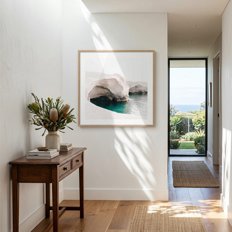 A framed art print depicting a coastal rock formation over turquoise water, hung on a white wall in a modern, sunlit hallway with a wooden console table and plants.